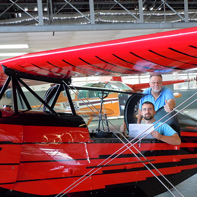 Two men in a red biplane with one man waving.